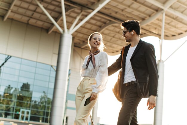 Hombre alegre con gafas de sol de traje negro abraza a su novia con blusa blanca y pantalones beige Atractiva mujer y chico de buen humor caminan cerca del aeropuerto