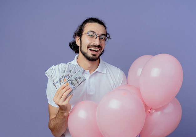 Foto gratuita hombre alegre con gafas sosteniendo dinero en efectivo y globos aislados sobre fondo púrpura