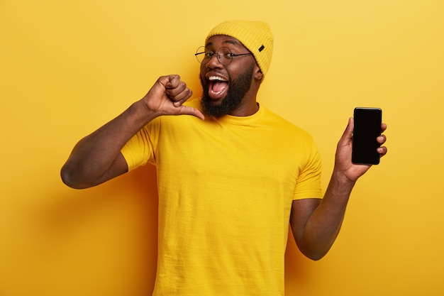 Un hombre alegre y guapo se señala a sí mismo, se siente orgulloso de desarrollar una nueva aplicación, muestra la pantalla del teléfono móvil