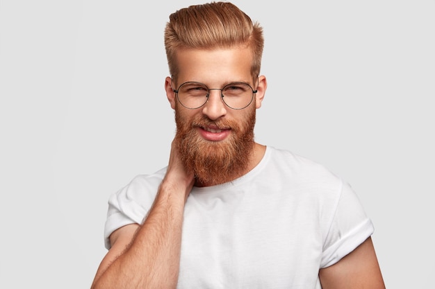Foto gratuita hombre alegre de jengibre con barba y bigote espesos, mira misteriosamente a través de gafas, viste camisa casual