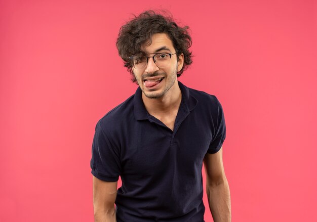 Hombre alegre joven en camisa negra con gafas ópticas pega la lengua fuera aislado en la pared rosa