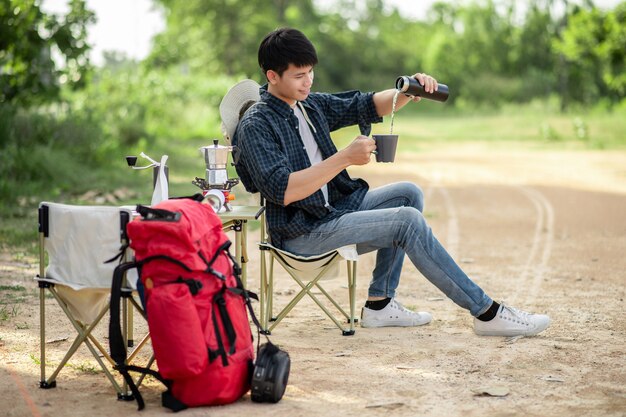 Hombre alegre joven mochilero sentado en la parte delantera de la carpa en el bosque con juego de café y haciendo molinillo de café recién hecho durante el viaje de campamento en las vacaciones de verano