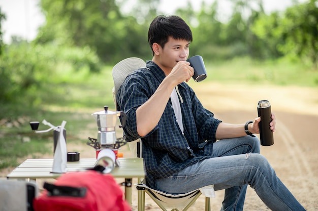 Hombre alegre joven mochilero sentado en la parte delantera de la carpa en el bosque con juego de café y haciendo molinillo de café recién hecho durante el viaje de campamento en las vacaciones de verano