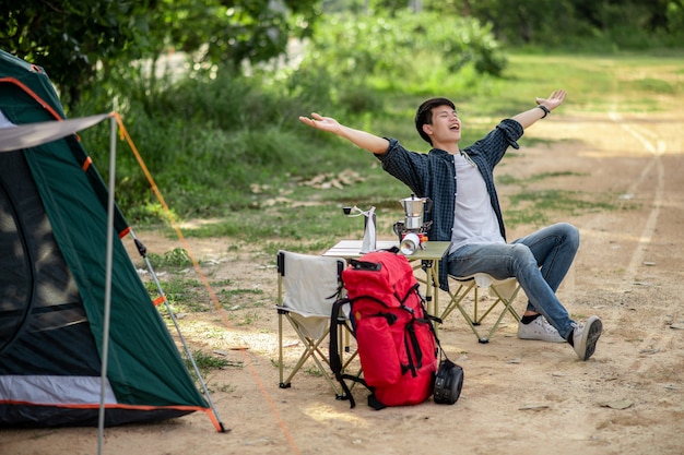 Hombre alegre joven viajero sentado en la parte delantera de la carpa en el bosque con juego de café y haciendo molinillo de café fresco durante el viaje de campamento en las vacaciones de verano