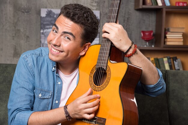 Hombre alegre mostrando una hermosa guitarra y sentado en el sofá. Foto de alta calidad