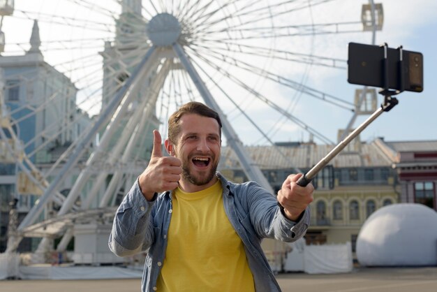 Hombre alegre mostrando pulgar arriba gesto mientras toma selfie delante de la noria