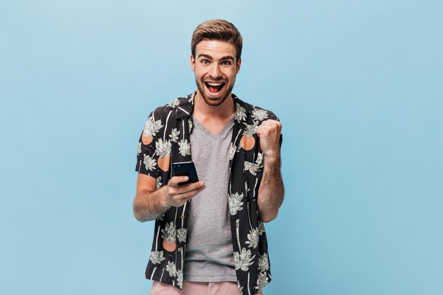 Hombre alegre con ojos azules en ropa fresca de moda mirando a la cámara se regocija y posando con el teléfono inteligente en un contexto aislado