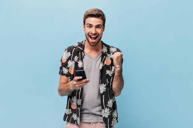 Hombre alegre con ojos azules en ropa fresca de moda mirando a la cámara se regocija y posando con el teléfono inteligente en un contexto aislado