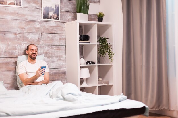 Hombre alegre en pijama riendo mientras ve un video divertido en el teléfono inteligente por la noche en una cama cómoda.