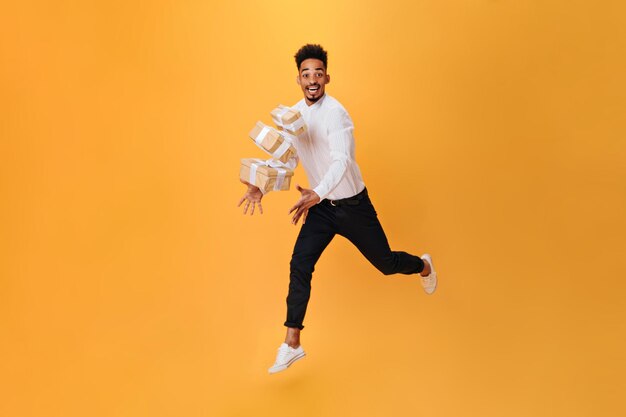 Hombre alegre salta sobre fondo naranja y lanza regalos Hombre de pelo oscuro con camisa blanca y pantalón negro posando felizmente en aislado