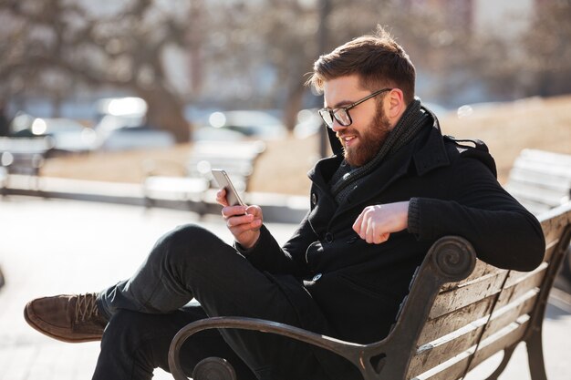 Hombre alegre sentado y usando el teléfono inteligente en la ciudad