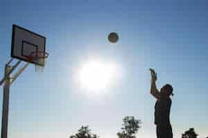 Foto gratuita hombre alto jugando baloncesto solo en un día soleado al aire libre en el campo de deportes. hombre lanzando baloncesto a la canasta contra el fondo del cielo cenital. estilo de vida activo, deporte, concepto de motivación.