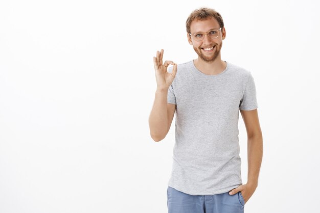 Hombre amable y servicial inteligente sin afeitar con gafas transparentes y atuendo casual sosteniendo la mano en el bolsillo sonriendo casualmente seguro y mostrando un gesto bien o bien sobre una pared blanca