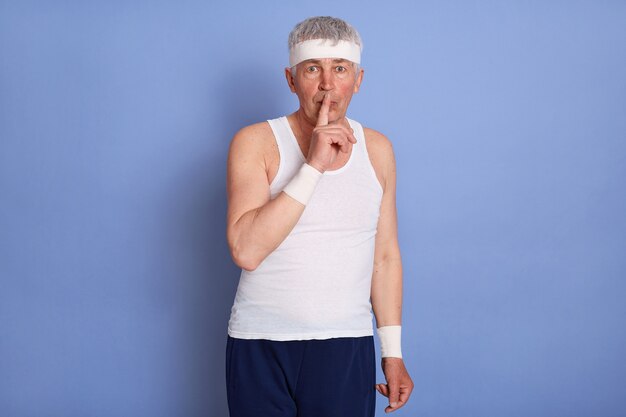 Hombre anciano con estilo atractivo con camiseta blanca sin mangas, haciendo un gesto de silencio, sosteniendo el dedo índice en sus labios, pidiendo mantener su secreto, posando.