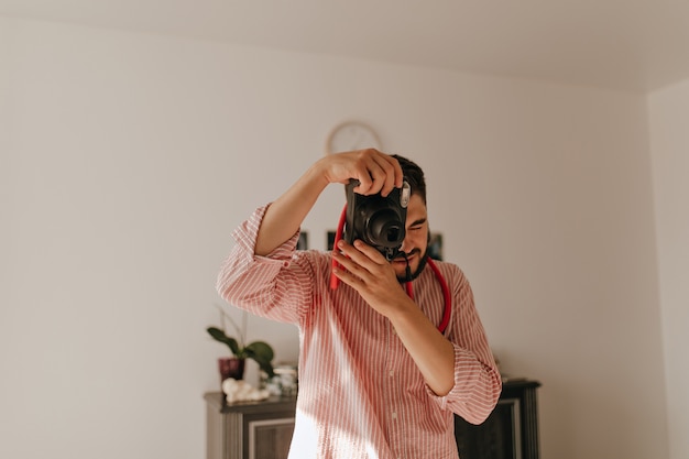 El hombre con el anillo en el dedo hace una foto en la cámara. Instantánea de chico moreno con traje de rayas en apartamento espacioso.