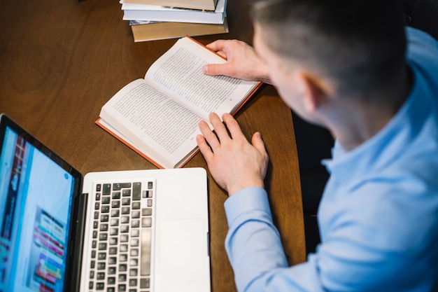 Hombre anónimo que lee el libro cerca de la computadora portátil