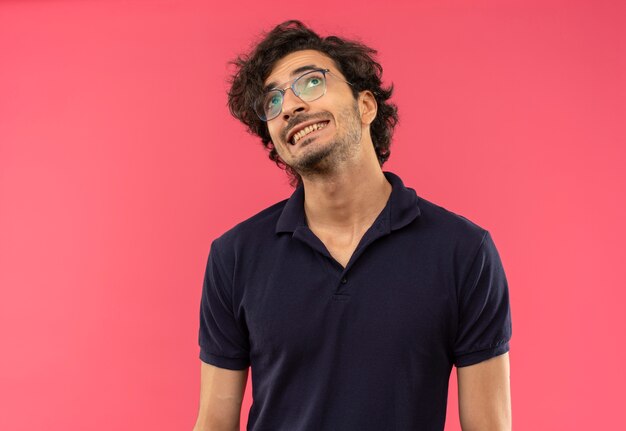 Hombre ansioso joven en camisa negra con gafas ópticas aprieta los dientes y mira hacia arriba aislado en la pared rosa