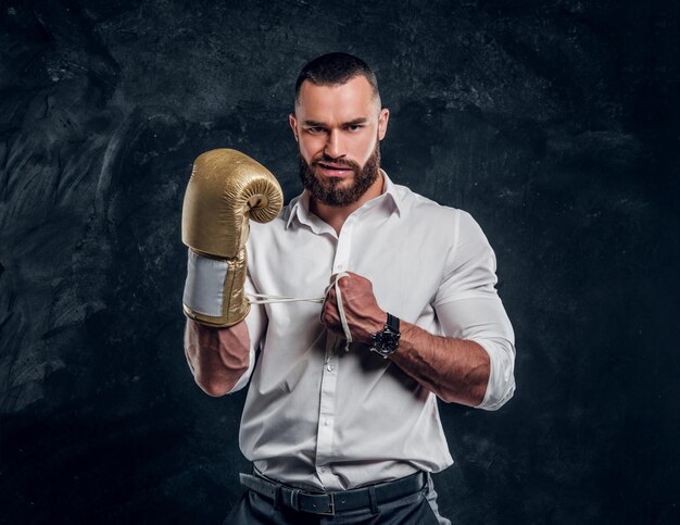 Un hombre apuesto y descarado con camisa blanca y guante de boxeo dorado está parado en un estudio oscuro.