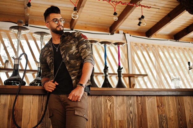 Hombre árabe de barba elegante con gafas y chaqueta militar fumando narguile en el bar de la calle Modelo árabe descansando