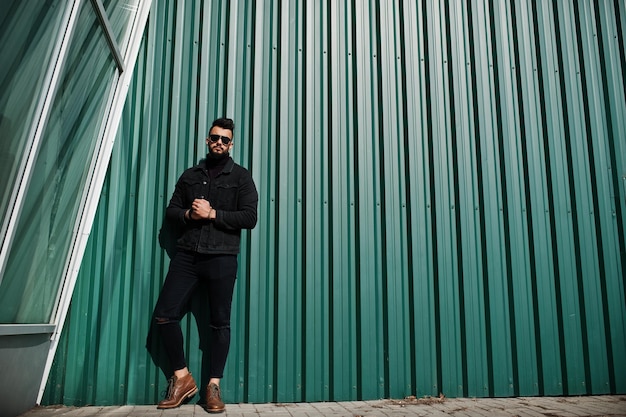 Hombre árabe de barba rica en moda con chaqueta de jeans negros y gafas de sol posados contra la pared verde con ventanas Elegante modelo árabe exitoso y de moda