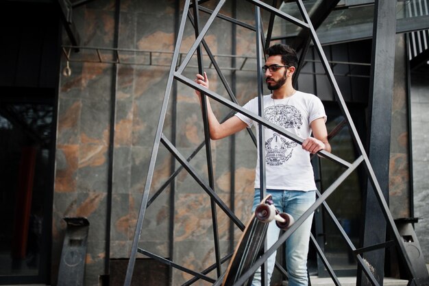 Hombre árabe de estilo callejero en anteojos con longboard posado dentro de la construcción de pirámide de metal