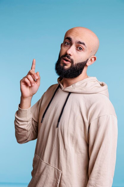 Hombre árabe inspirado que tiene una idea y levanta el dedo índice mientras mira la cámara. Joven calvo barbudo con expresión facial emocionada apuntando hacia arriba retrato de estudio