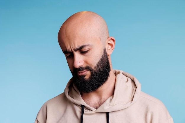 Foto gratuita hombre árabe pensativo inclinando la cabeza hacia abajo y mirando hacia otro lado. persona joven con barba calva pensando con expresión facial incierta y posando casualmente en el estudio sobre fondo azul