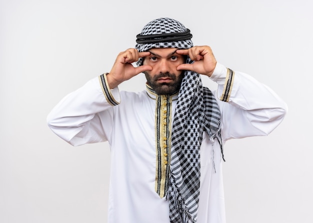 Hombre árabe en ropa tradicional abriendo los ojos con los dedos tratando de ver mejor parado sobre la pared blanca