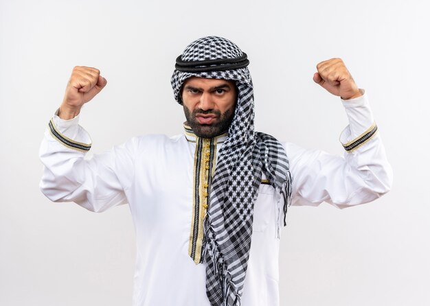 Hombre árabe en ropa tradicional apretando el puño levantando las manos con cara seria de pie sobre la pared blanca