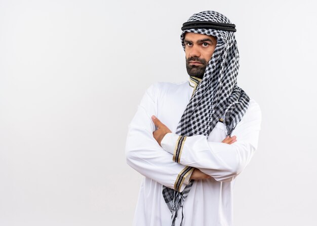 Hombre árabe en ropa tradicional con los brazos cruzados sobre el pecho mirando a un lado con cara seria de pie sobre la pared blanca