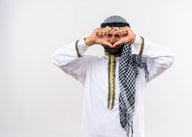 Hombre árabe en ropa tradicional haciendo gesto de corazón con los dedos mirando a través de los dedos sonriendo de pie sobre la pared blanca