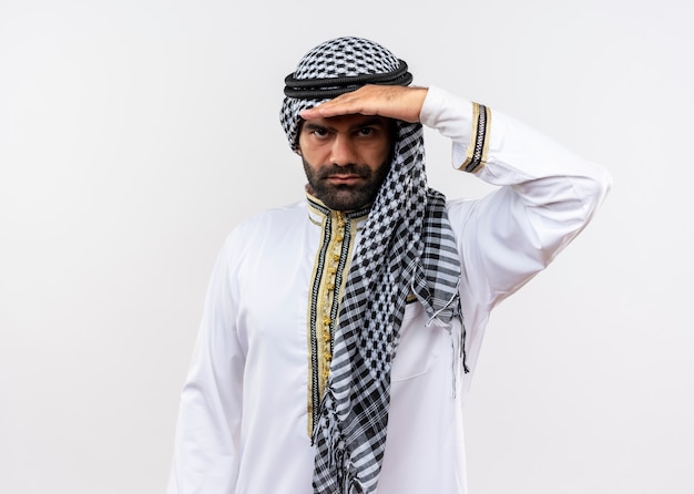 Hombre árabe en ropa tradicional mirando lejos con las manos sobre la cabeza de pie sobre una pared blanca