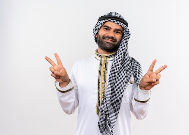 Foto gratuita hombre árabe en ropa tradicional sonriendo positivo y feliz mostrando el signo de la victoria con ambas manos de pie sobre la pared blanca