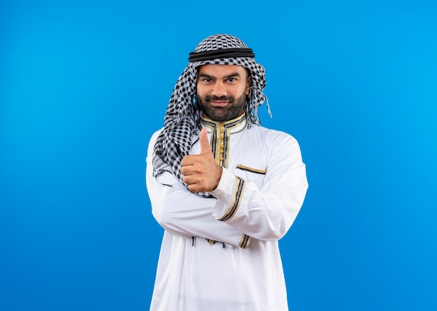 Foto gratuita hombre árabe en ropa tradicional con una sonrisa en la cara mostrando los pulgares para arriba de pie sobre la pared azul