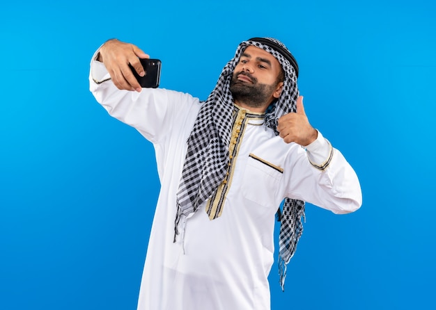 Foto gratuita hombre árabe en ropa tradicional tomando selfie con smartphone mostrando los pulgares para arriba de pie sobre la pared azul