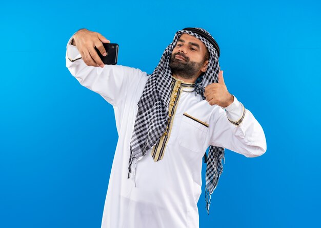 Hombre árabe en ropa tradicional tomando selfie con smartphone mostrando los pulgares para arriba de pie sobre la pared azul