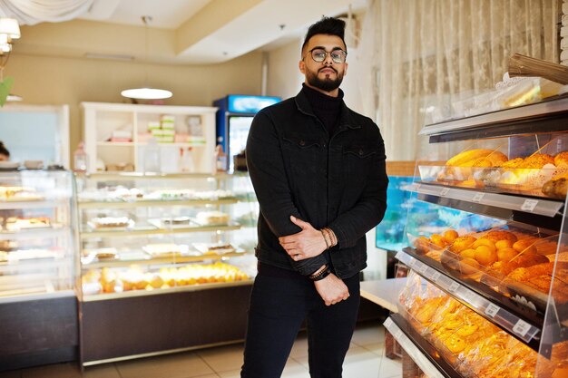 El hombre árabe usa una chaqueta de jeans negros y anteojos en un café, elige una panadería, un modelo árabe elegante y de moda.