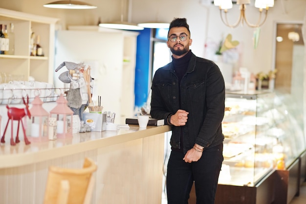 El hombre árabe usa una chaqueta negra de jeans y anteojos en un café, bebe café en el bar, un modelo árabe elegante y de moda.