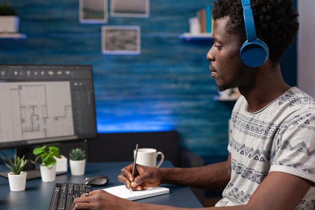 Hombre arquitecto afroamericano con auriculares analizando el plan del modelo de edificio desarrollando el boceto del edificio de rascacielos. Joven ingeniero que trabaja de forma remota desde casa en el desarrollo del concepto de construcción de edificios