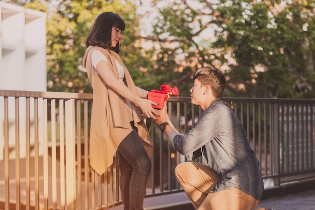 Hombre arrodillado dando a su novia rosas y un regalo rojo