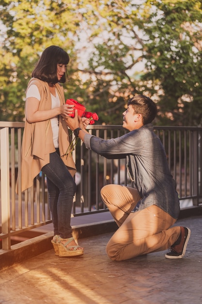 Foto gratuita hombre arrodillado dando a su novia rosas y un regalo rojo