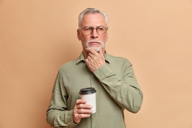 El hombre arrugado pensativo se para en pose pensativa se frota la barbilla e intenta que su mente contemple algo mientras toma un café, usa gafas ópticas y camisa formal.