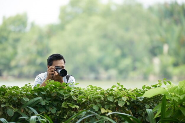 Hombre asiático con cámara profesional mirando por encima del seto verde en el parque y tomando fotos