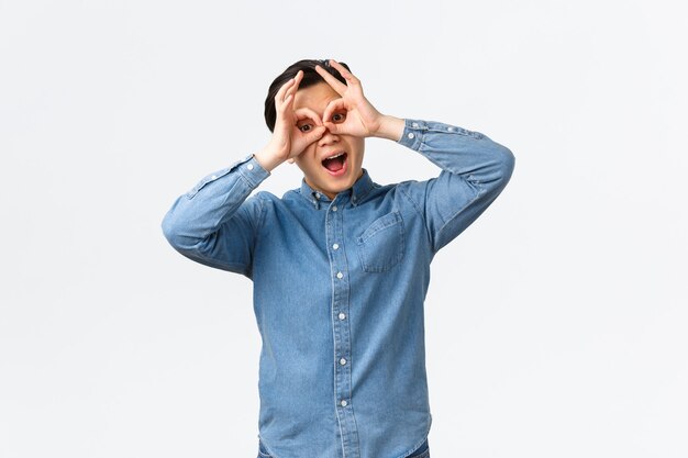 Hombre asiático divertido y alegre, juguetón haciendo muecas, mostrando gafas falsas con las manos sobre los ojos, burlándose de alguien, jugando, viendo algo emocionante, de pie con un fondo blanco.