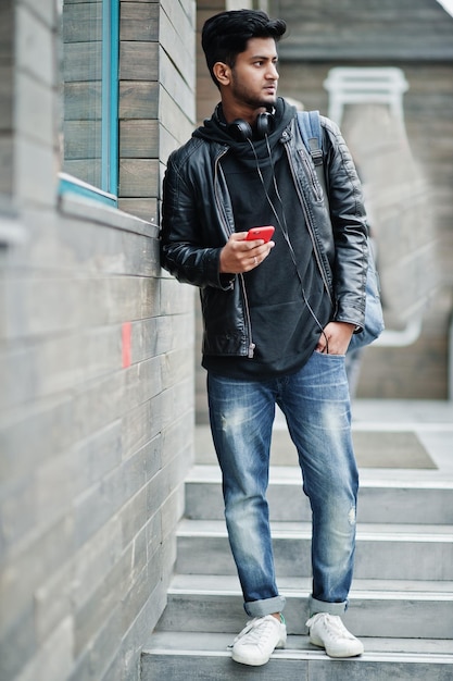 Hombre asiático elegante e informal con auriculares de chaqueta de cuero negro con teléfono móvil rojo en las manos posado en la calle