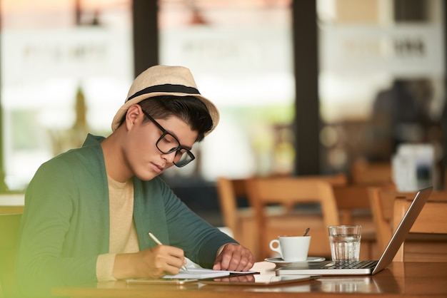 Hombre asiático elegante joven sentado en la cafetería con ordenador portátil y escribir en el cuaderno