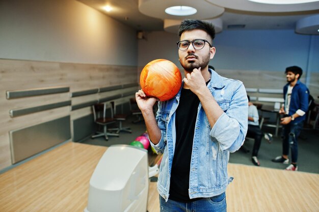 Hombre asiático con estilo en chaqueta de jeans y gafas de pie en la bolera con la pelota en la mano