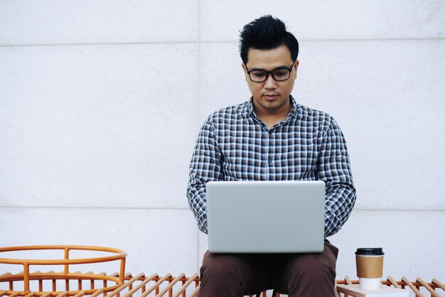 Hombre asiático en gafas sentado en el banco al aire libre y trabajando en la computadora portátil