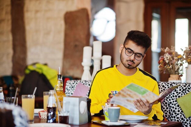 Hombre asiático con gafas y sudadera amarilla sentado en un café y leyendo el menú