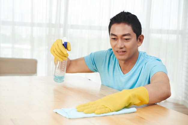 Hombre asiático en guantes de goma rociando mesa y limpieza con un paño en casa